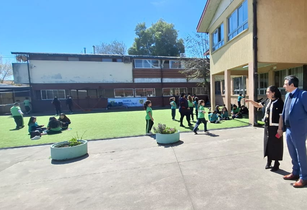 Fotografía del patio de una escuela, donde hay niñas y niños corriendo y a la derecha, una mujer mostrando el lugar a un hombre