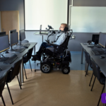 Foto de una persona en silla de ruedas en una sala de universidad llena de compitadores