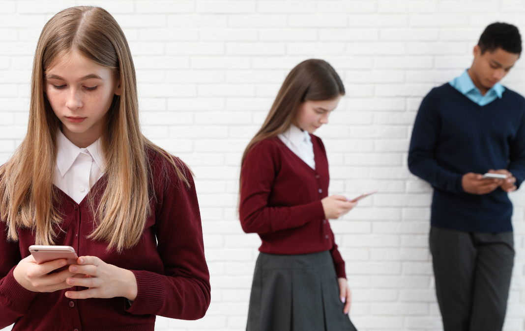 Tres adolescentes, dos mujeres y un hombre con uniforme escolar, mirando sus teléfonos celulares