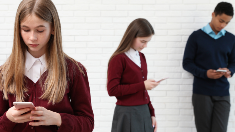 Tres adolescentes, dos mujeres y un hombre con uniforme escolar, mirando sus teléfonos celulares