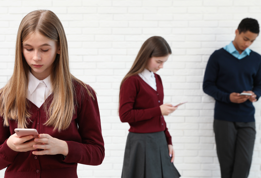 Tres adolescentes, dos mujeres y un hombre con uniforme escolar, mirando sus teléfonos celulares