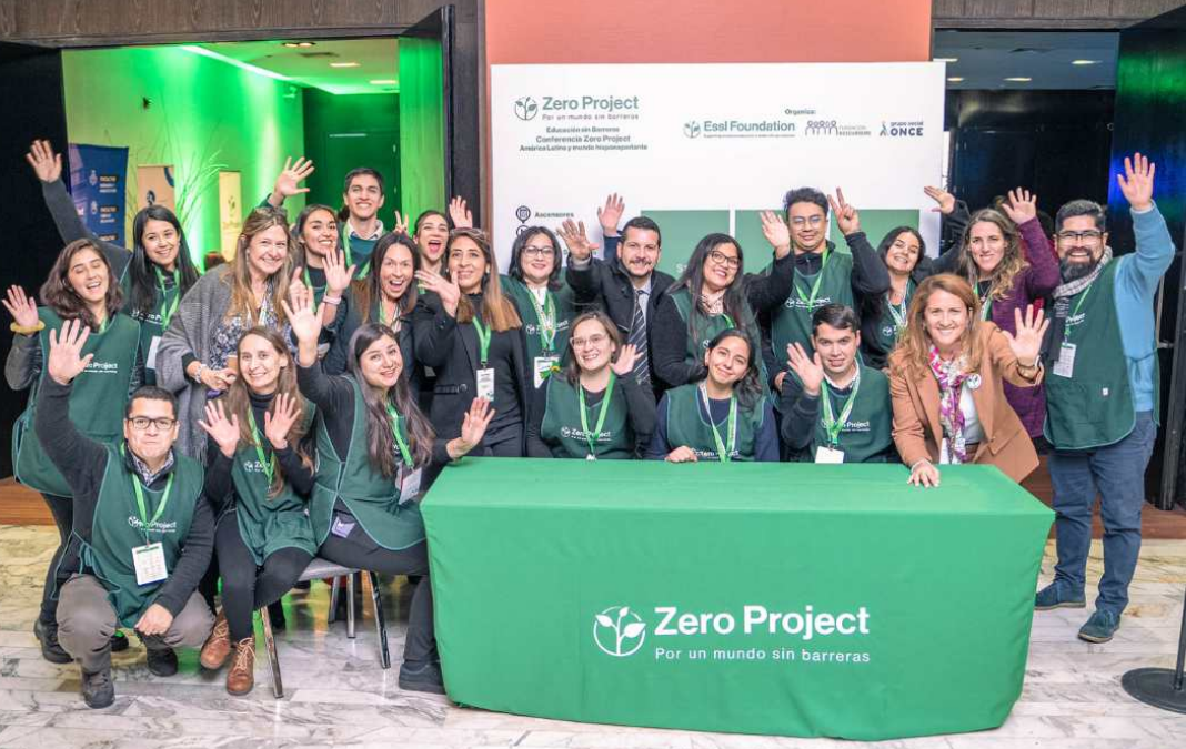 Imagen de varias personas saludando a la cámara junto a una mesa con mantel verde que dice Zero Project