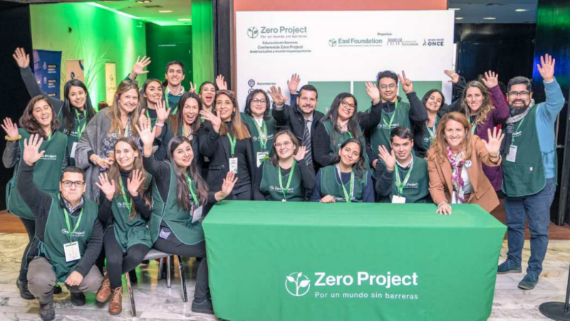 Imagen de varias personas saludando a la cámara junto a una mesa con mantel verde que dice Zero Project