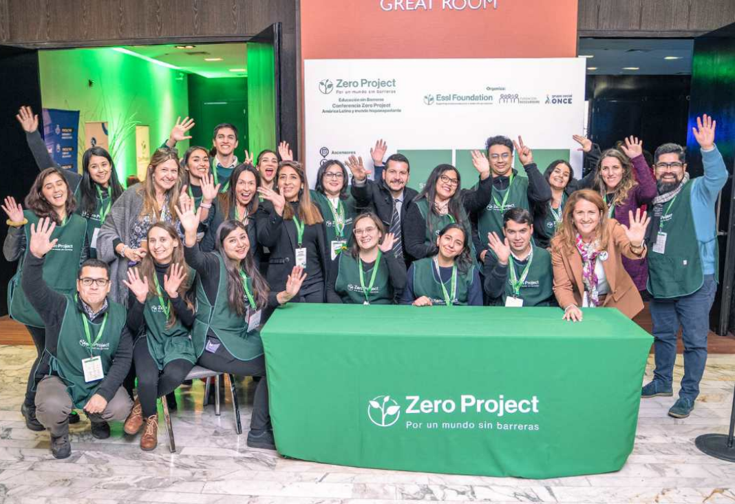 Imagen de varias personas saludando a la cámara junto a una mesa con mantel verde que dice Zero Project