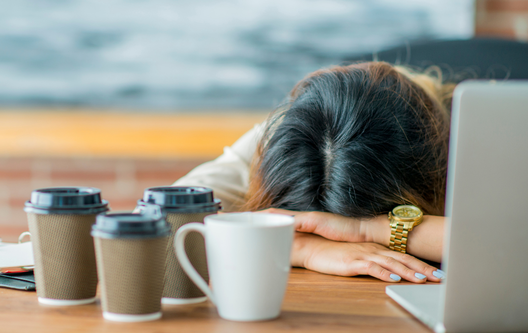Mujer acostada sobre una mesa y rodeada de tazas de café y un computador