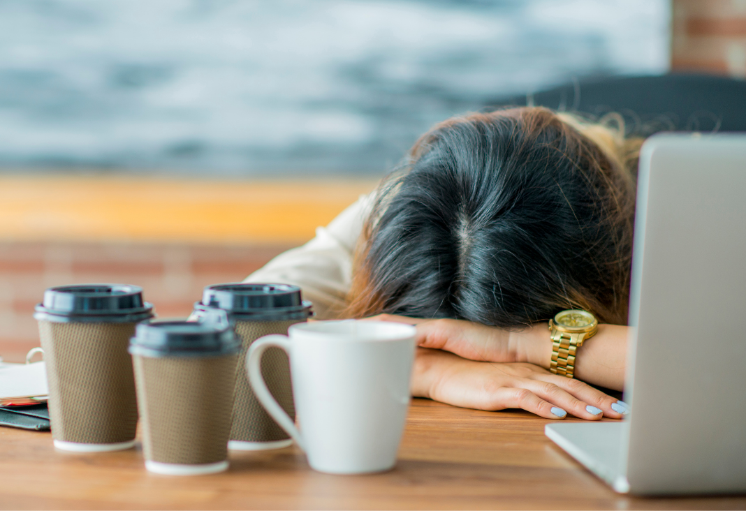 Mujer acostada sobre una mesa y rodeada de tazas de café y un computador