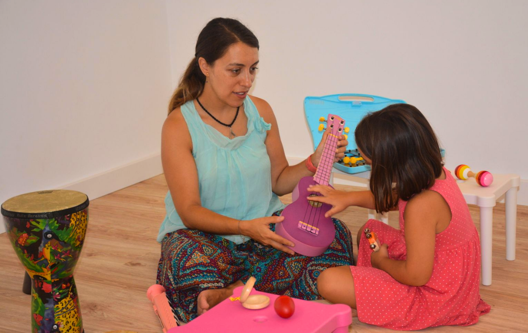 Mujer sentada en el piso junto a una niña y mostrándole un ukelele