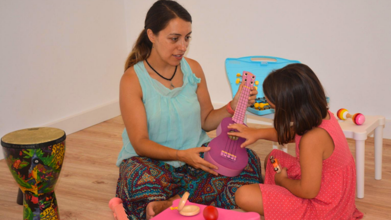 Mujer sentada en el piso junto a una niña y mostrándole un ukelele