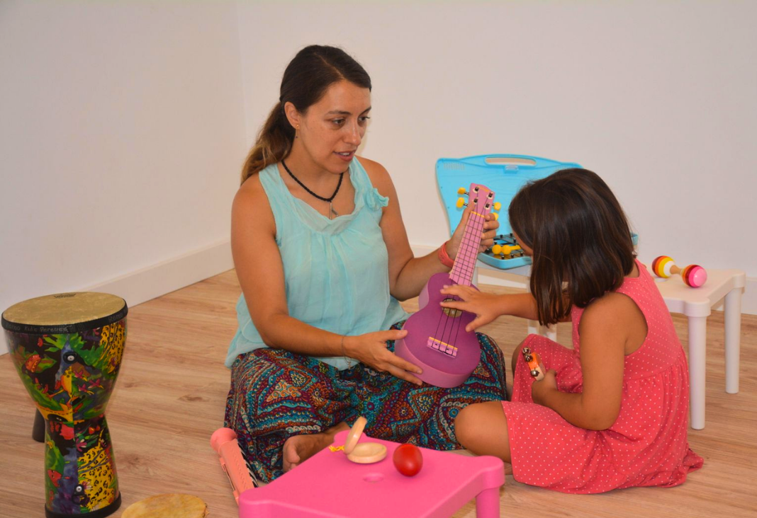 Mujer sentada en el piso junto a una niña y mostrándole un ukelele