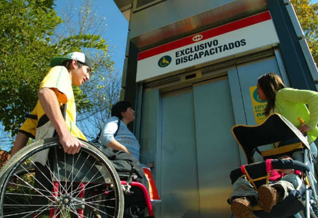 Tres personas esperando ascensor de metro: una señora, un hombre en silla de ruedas y una mujer con un coche