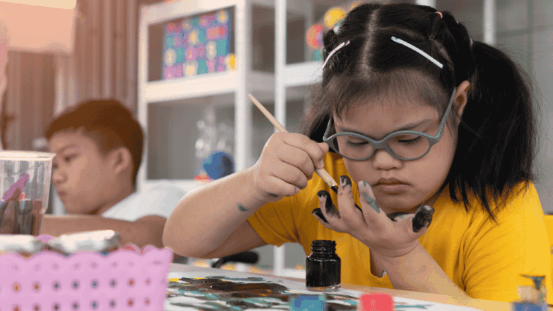 niña con síndrome de Down pintando y atrás otro niño
