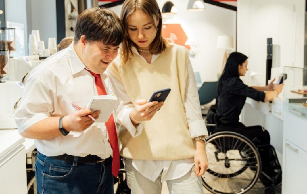 un hombre con sindrome de down junta a una colega de trabajo mirando un celular y atrás una mujer en silla de ruedas frente a un escritorio
