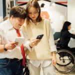 un hombre con sindrome de down junta a una colega de trabajo mirando un celular y atrás una mujer en silla de ruedas frente a un escritorio