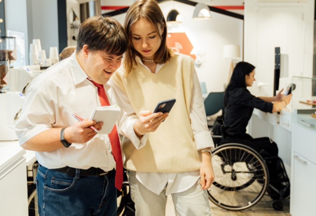 un hombre con sindrome de down junta a una colega de trabajo mirando un celular y atrás una mujer en silla de ruedas frente a un escritorio