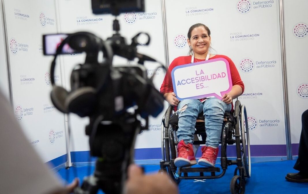 Una cámara de video y audifonos enfocando a una persona en silla de ruedas que sujeta un letrero que dice "La accesibilidad es..."
