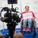 Una cámara de video y audifonos enfocando a una persona en silla de ruedas que sujeta un letrero que dice "La accesibilidad es..."