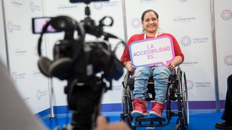 Una cámara de video y audifonos enfocando a una persona en silla de ruedas que sujeta un letrero que dice "La accesibilidad es..."
