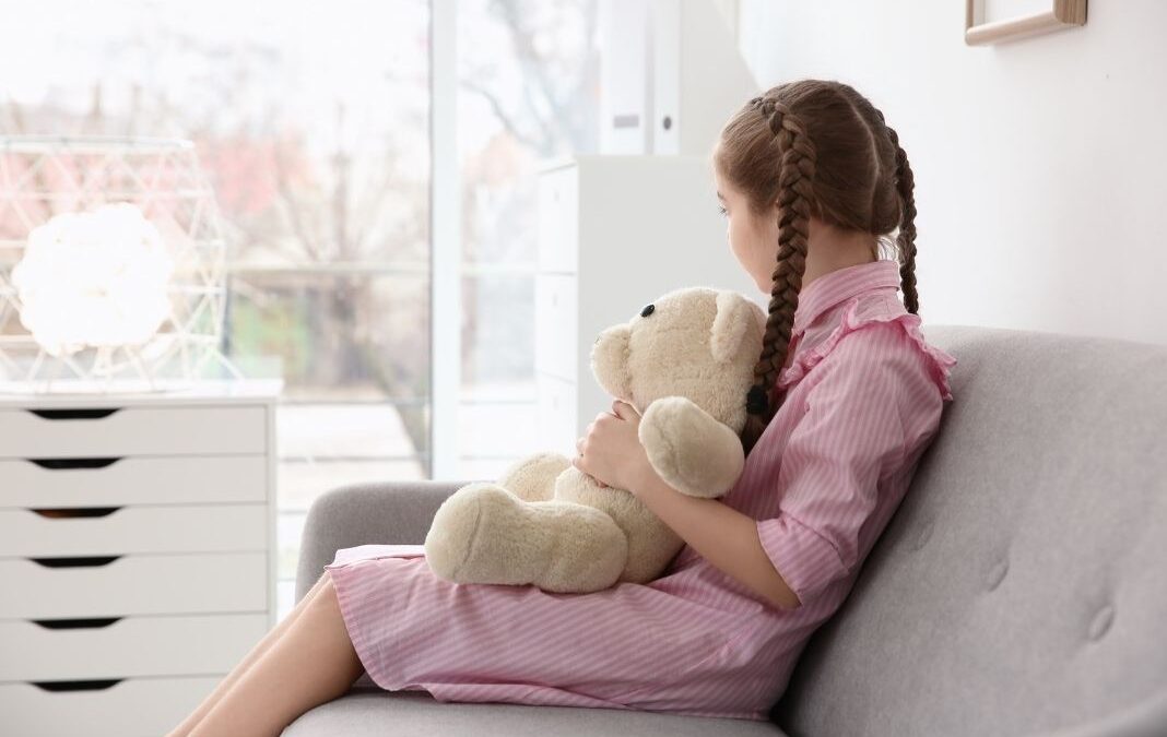 Niña sentada de lado mirando una ventana y con un oso de peluche en las piernas