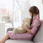 Niña sentada de lado mirando una ventana y con un oso de peluche en las piernas