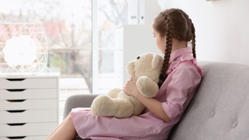 Niña sentada de lado mirando una ventana y con un oso de peluche en las piernas