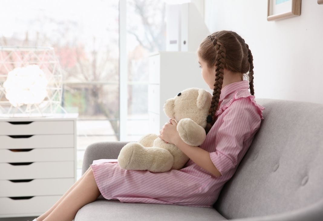 Niña sentada de lado mirando una ventana y con un oso de peluche en las piernas