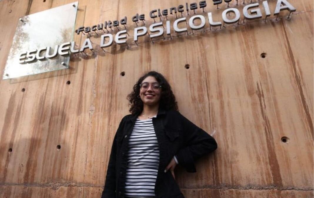 Joven mirando y sonriendo a la cámara, y arriba de ella letrero que dice "Escuela de Psicología"