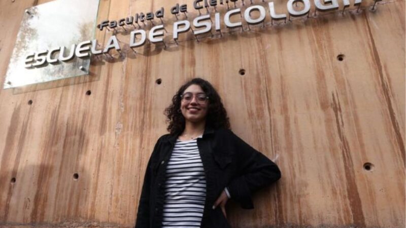 Joven mirando y sonriendo a la cámara, y arriba de ella letrero que dice "Escuela de Psicología"