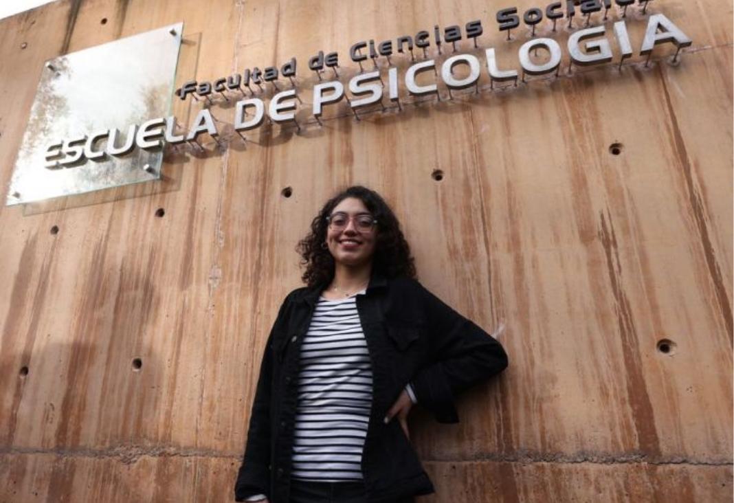 Joven mirando y sonriendo a la cámara, y arriba de ella letrero que dice "Escuela de Psicología"