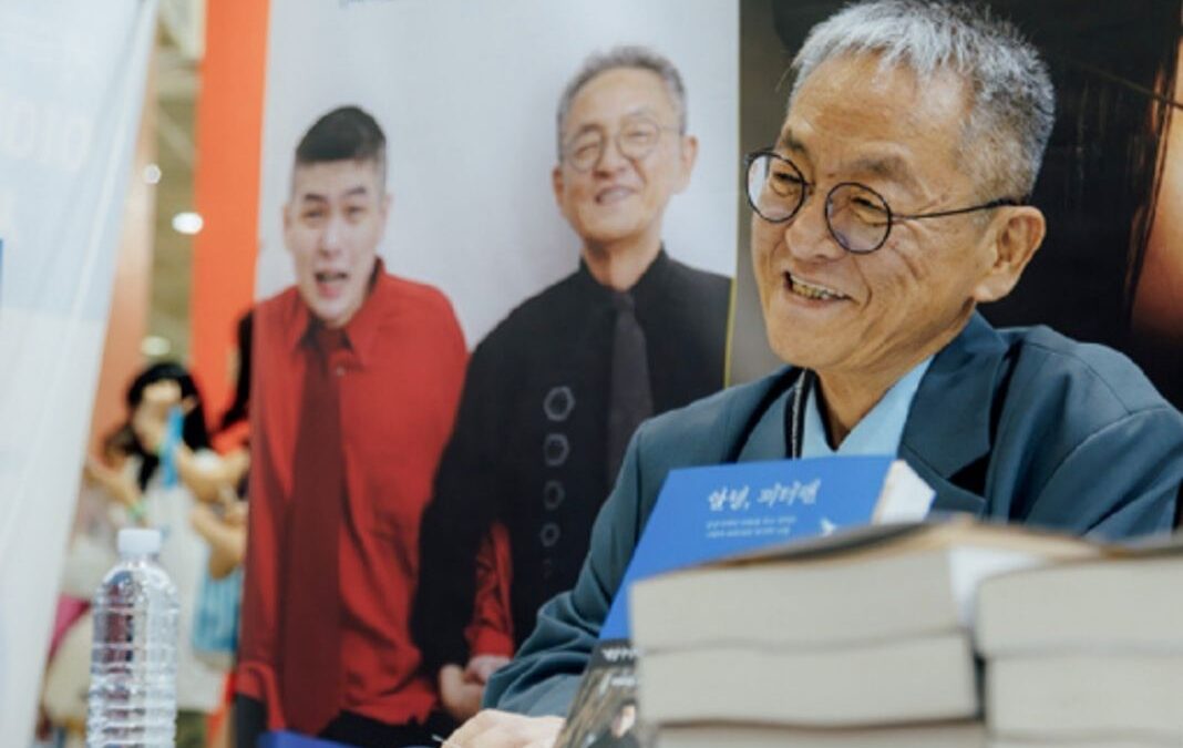 Hombre coreano sonriendo y libros sobre una mesa, atrás una foto de él y su hijo autista