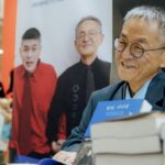 Hombre coreano sonriendo y libros sobre una mesa, atrás una foto de él y su hijo autista