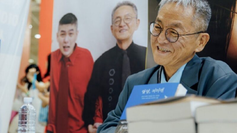 Hombre coreano sonriendo y libros sobre una mesa, atrás una foto de él y su hijo autista