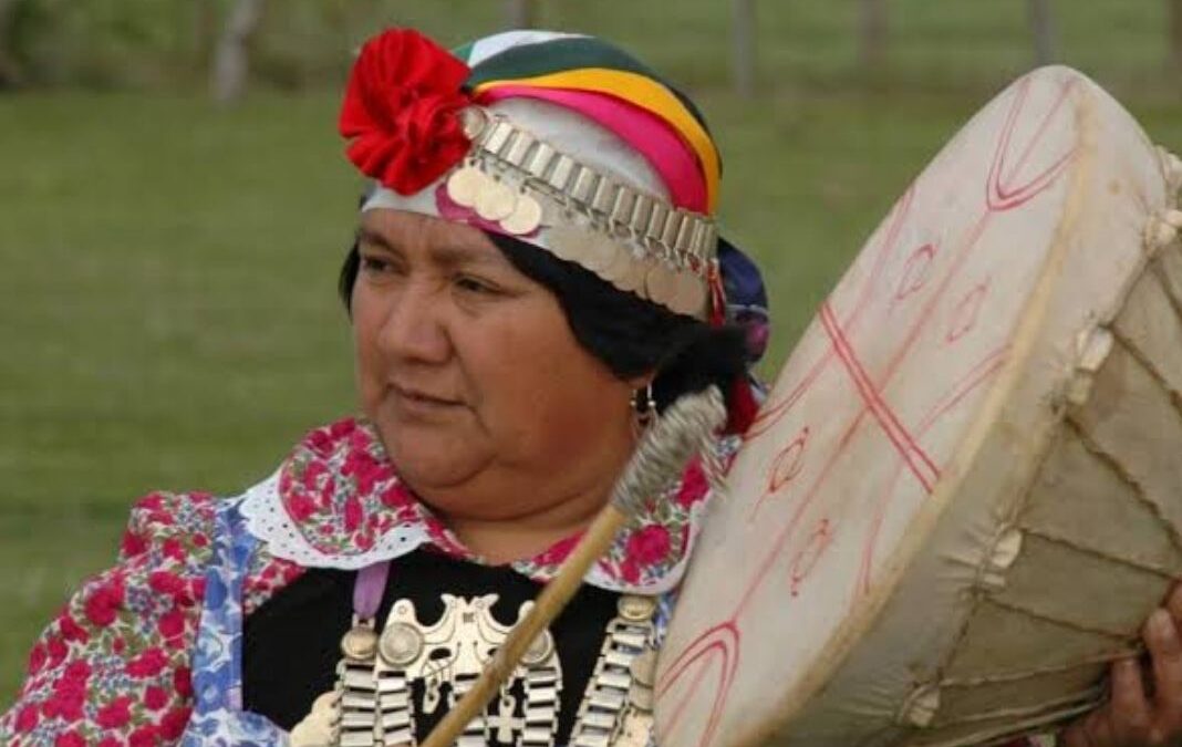 Mujer mapuche de perfil tocando un kultrün y con su vestimenta tradicional