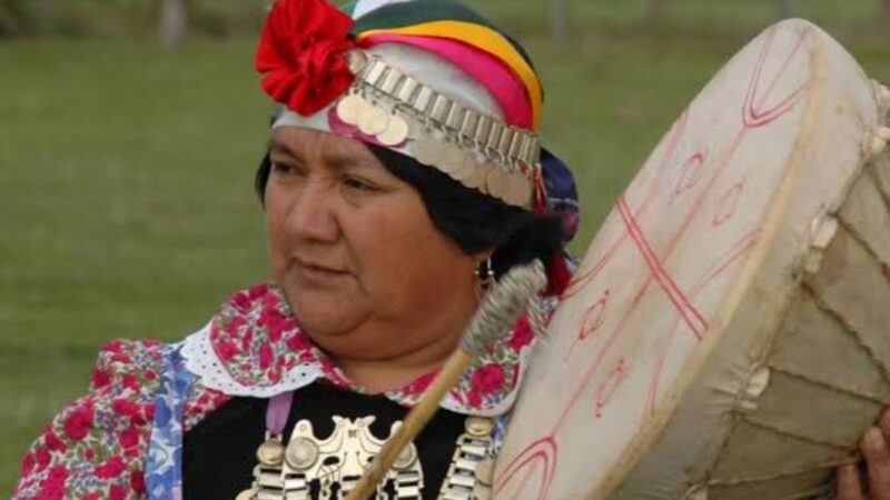 Mujer mapuche de perfil tocando un kultrün y con su vestimenta tradicional