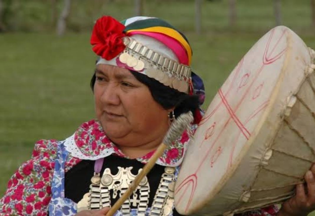 Mujer mapuche de perfil tocando un kultrün y con su vestimenta tradicional